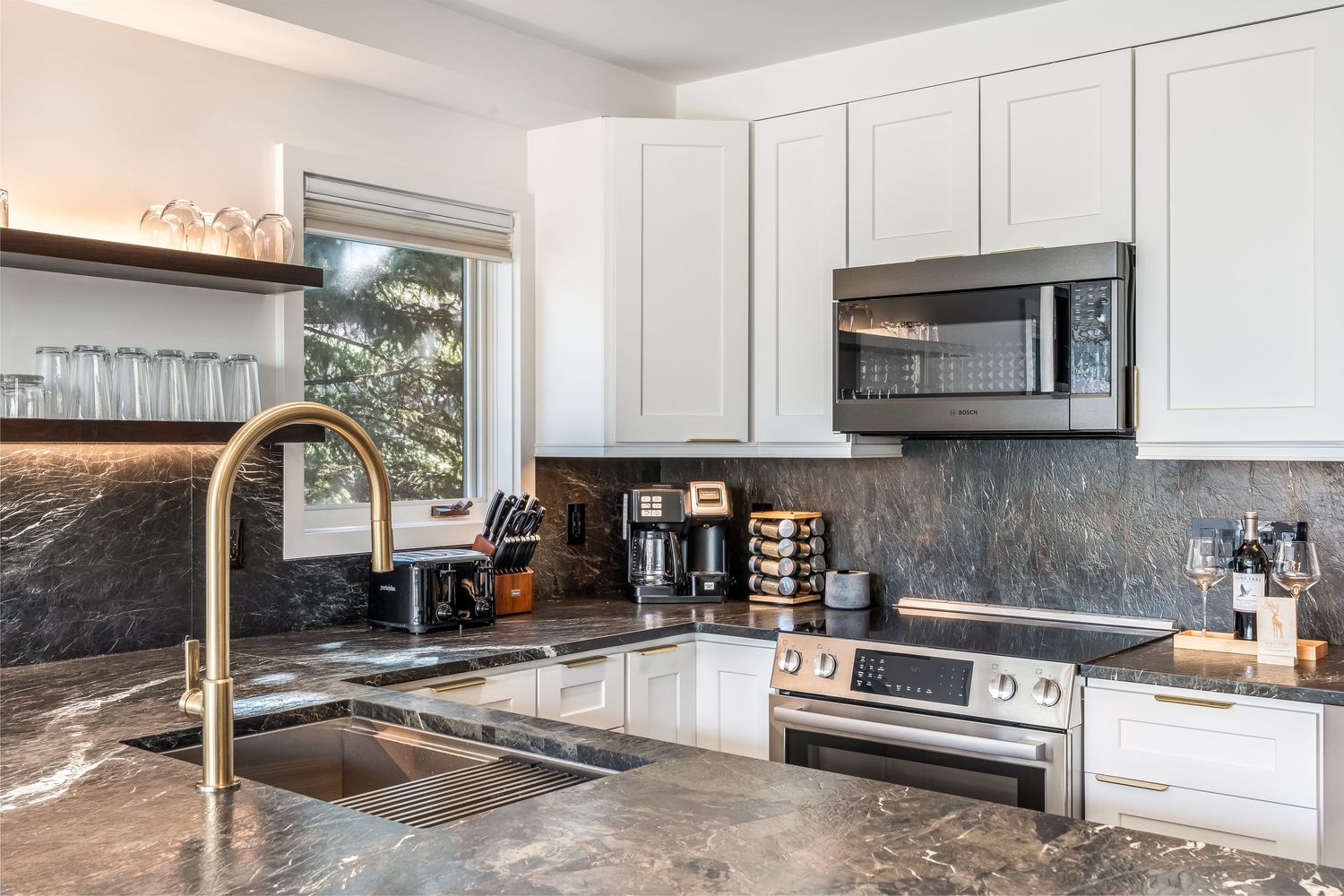 Modern kitchen with white cabinets, dark granite countertops, stainless steel appliances, and a gold faucet over a undermount sink.