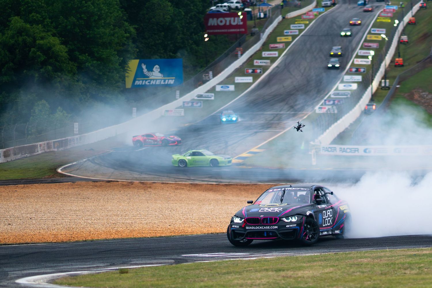 Race cars drift through smoky turns at a motorsport event on a scenic tree-lined circuit.