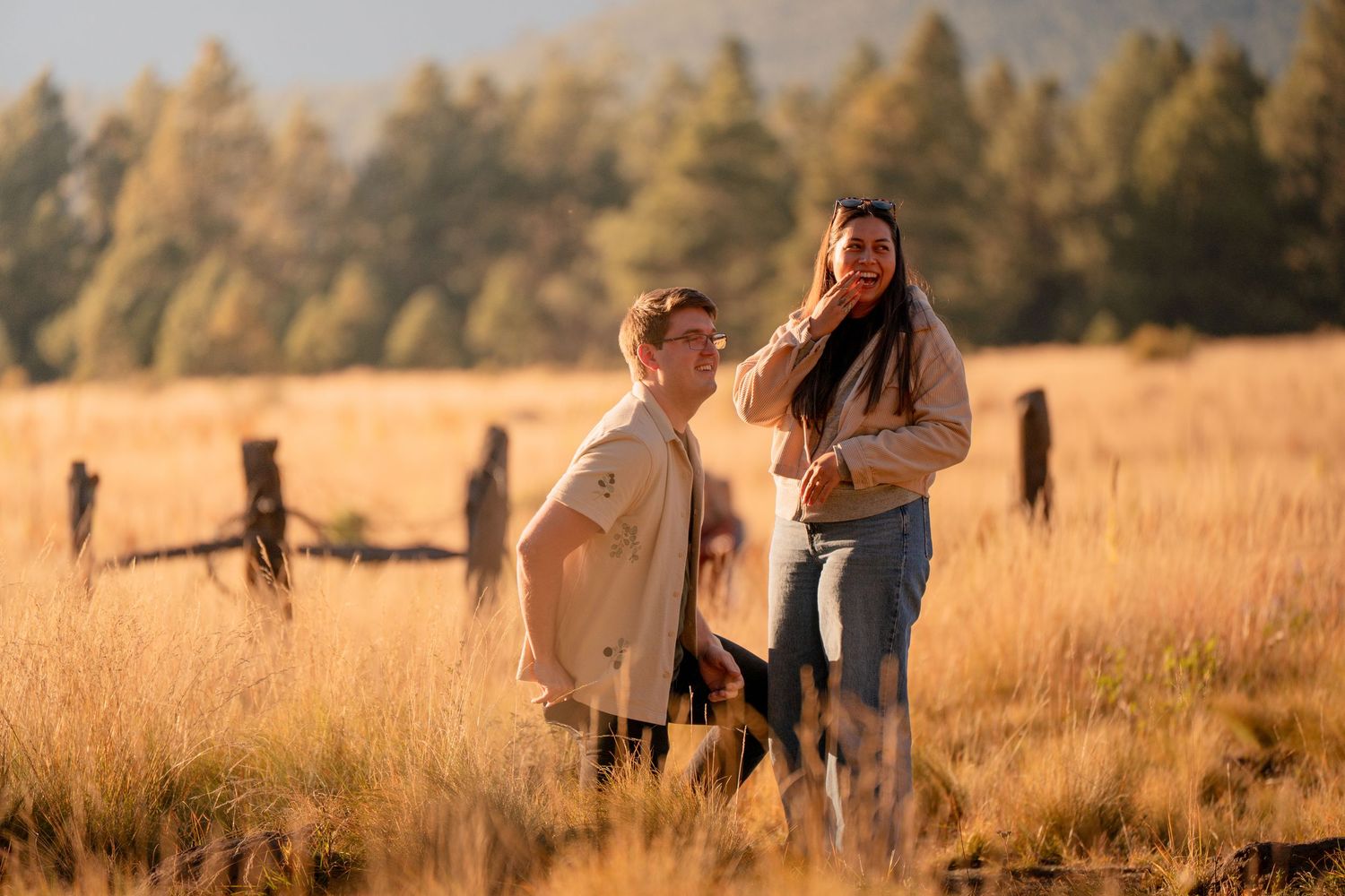 Smiling couple standing together in a golden grassy meadow with a rustic wooden fence and tall pine trees in the warm sunset light.