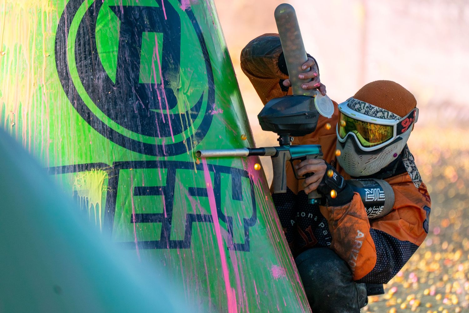 Paintball player in orange gear aims marker behind inflatable bunker during competitive speedball tournament.