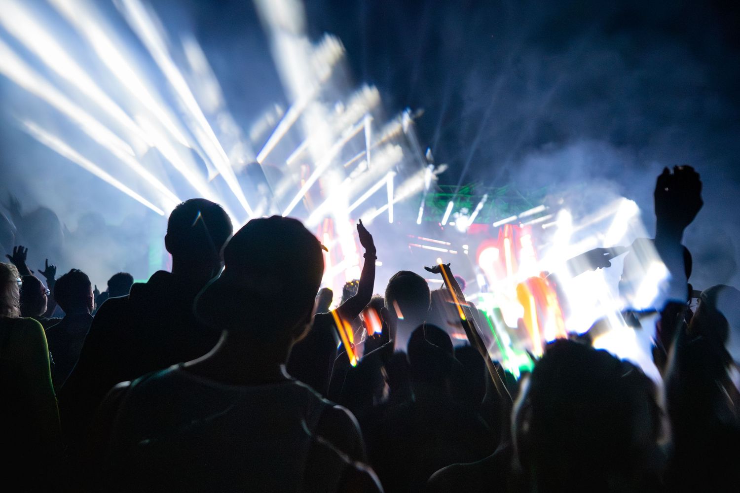 Crowd of silhouetted concert-goers with hands raised under bright stage lights and colorful laser beams at night.