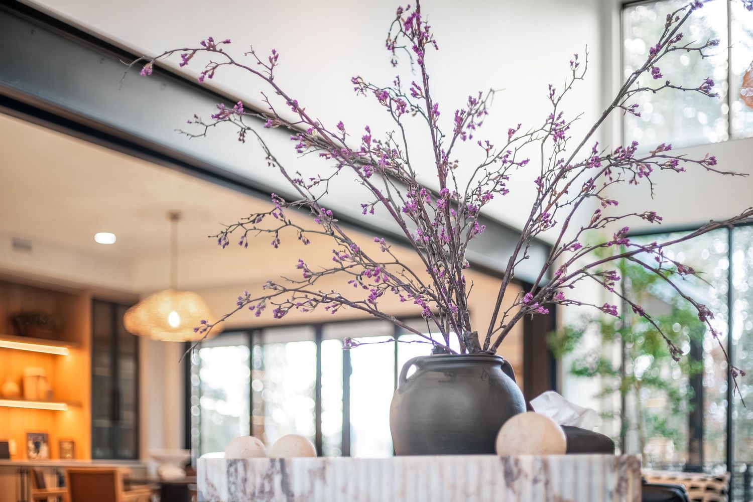 Modern living room interior with cherry blossom branches in tall black vases against floor-to-ceiling windows.