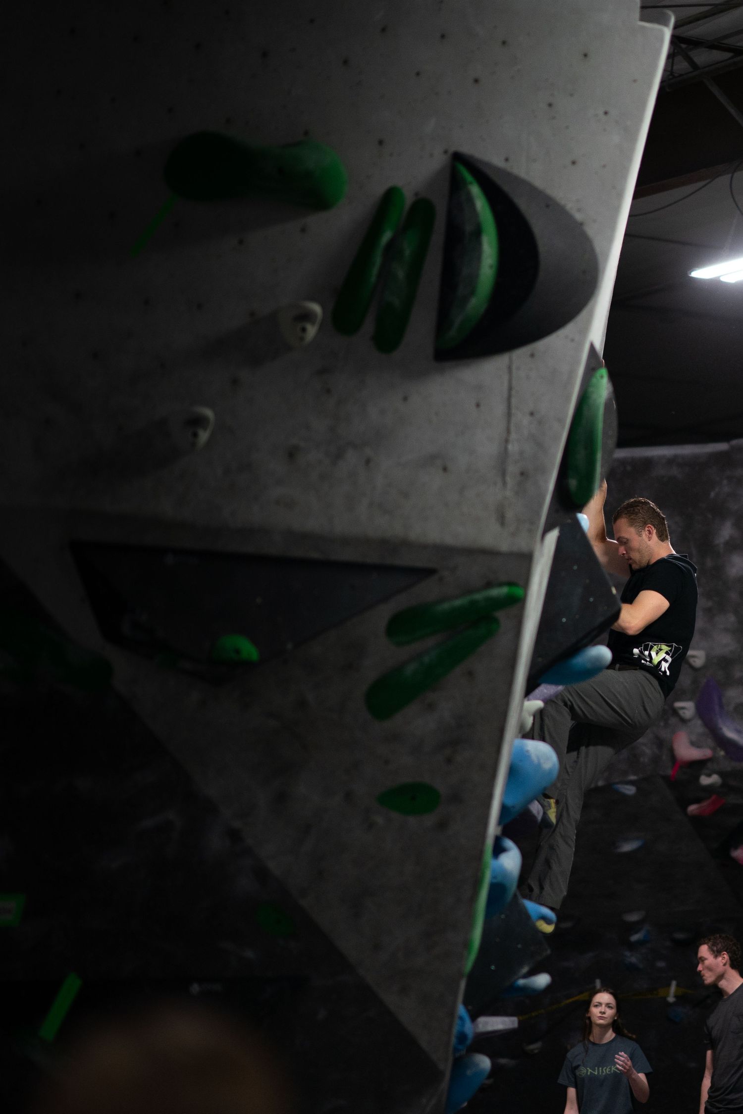 Climber in a black shirt scales an angled indoor bouldering wall dotted with green holds while two spectators watch from below.