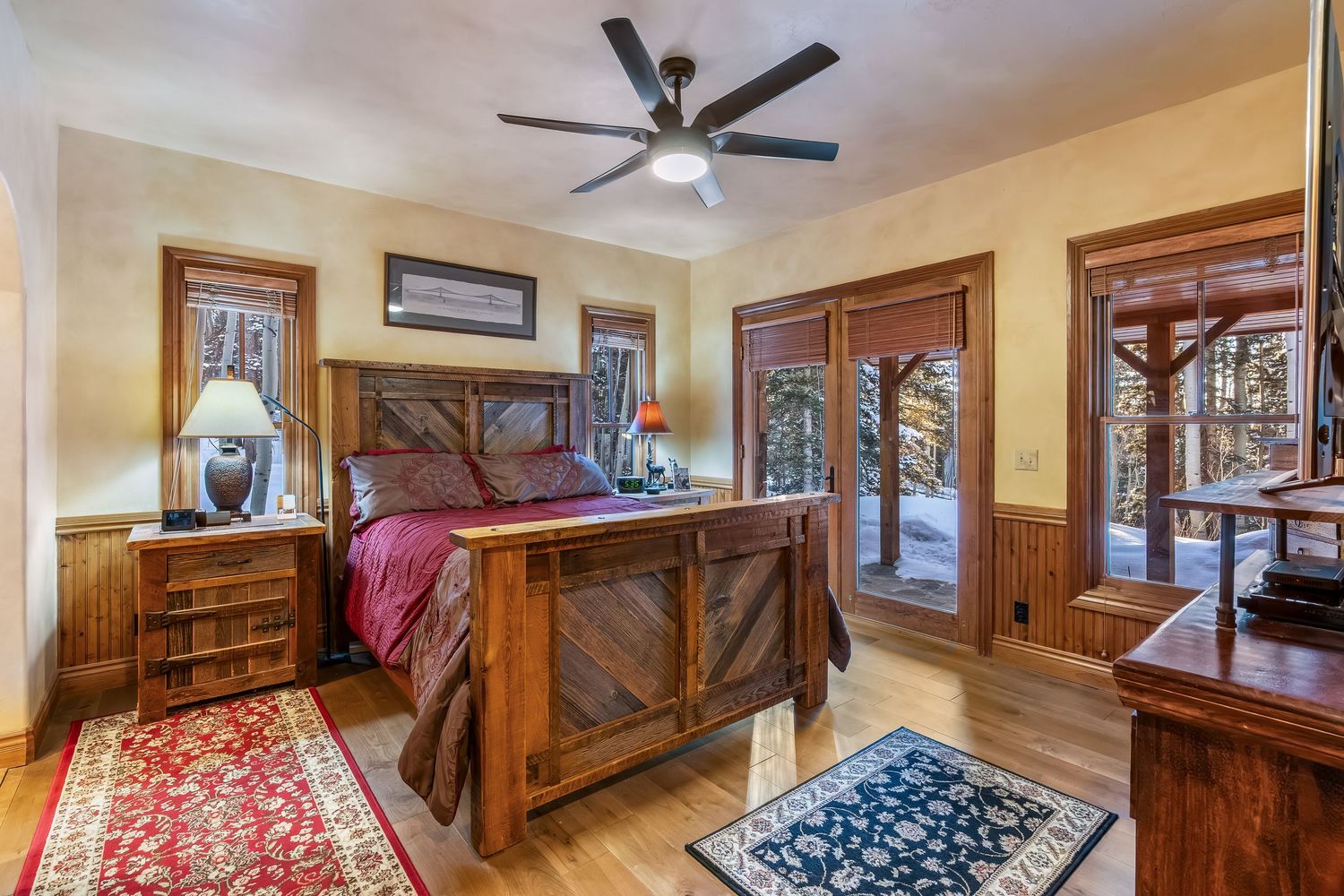 Rustic bedroom interior with wooden walls, ceiling fan, and natural light streaming through windows.