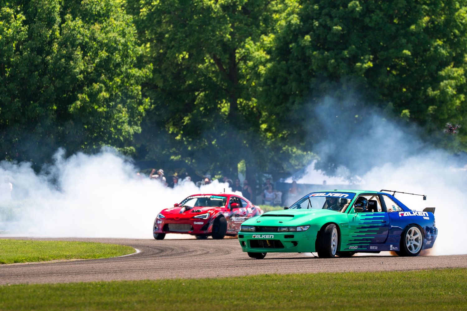Two drift cars racing on a track surrounded by smoke during a competitive drifting event on a sunny day.