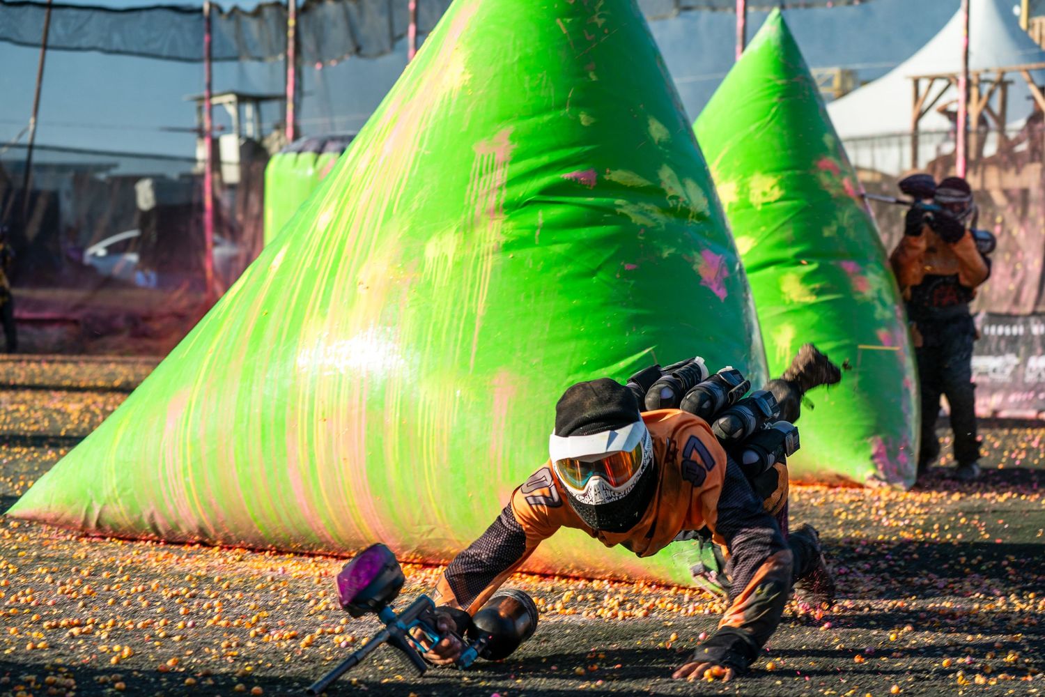 Paintball player diving behind green inflatable bunker during competitive speedball match on outdoor field.