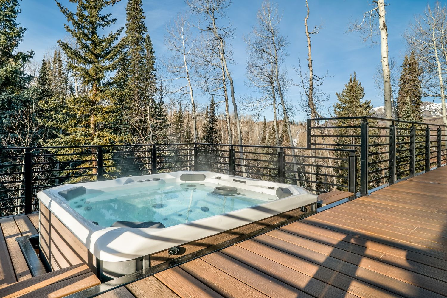 Panoramic deck view of a snowy mountain landscape with evergreen trees and a hot tub overlooking the scenery.