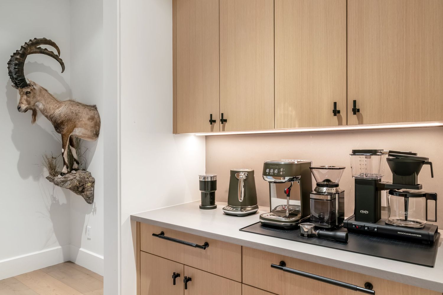 Sleek coffee station with mounted ibex head sculpture featuring espresso machines and coffee makers on wooden cabinets.