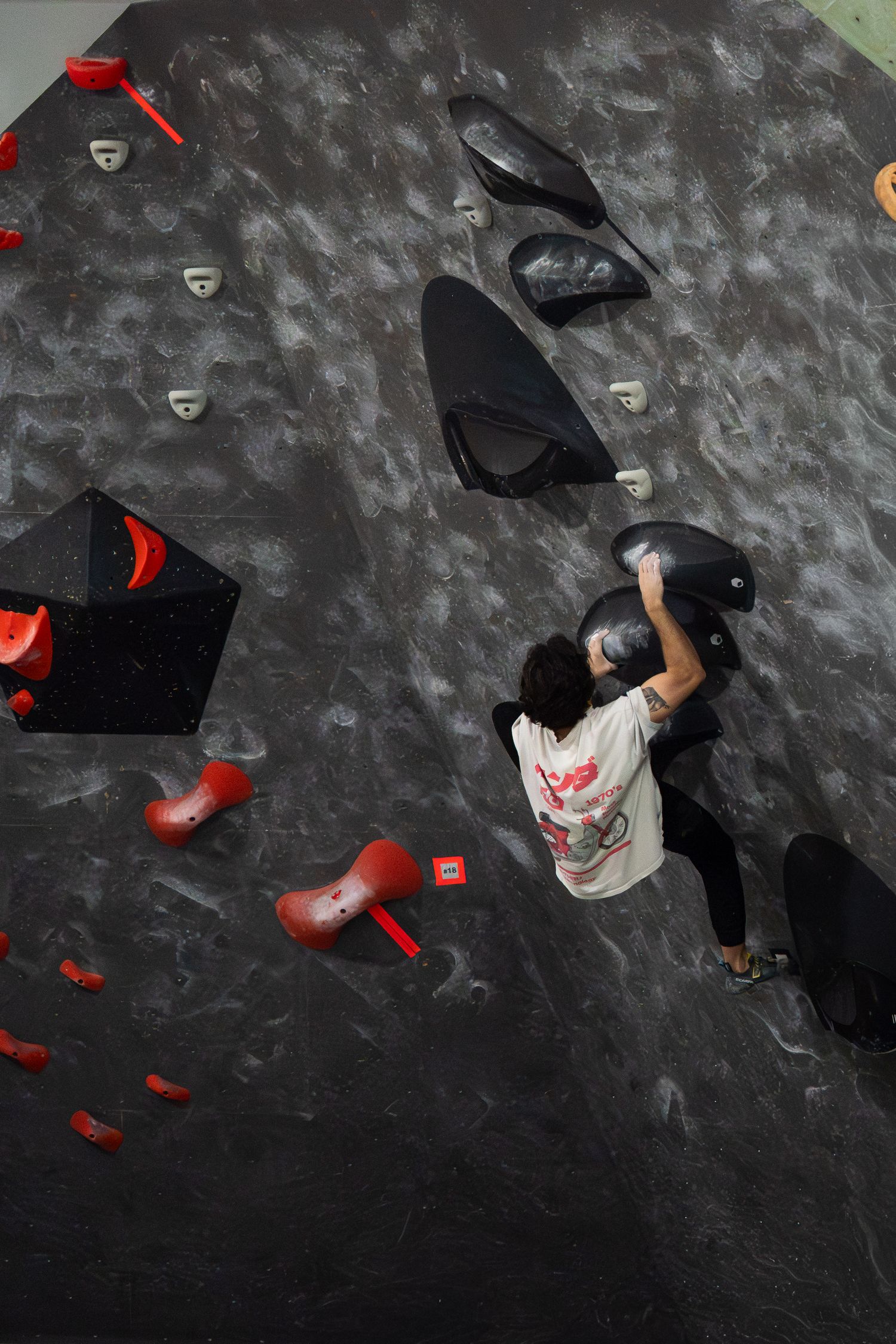 Climber in a white graphic tee reaches for a black hold on a dark indoor bouldering wall dotted with red and black volumes.