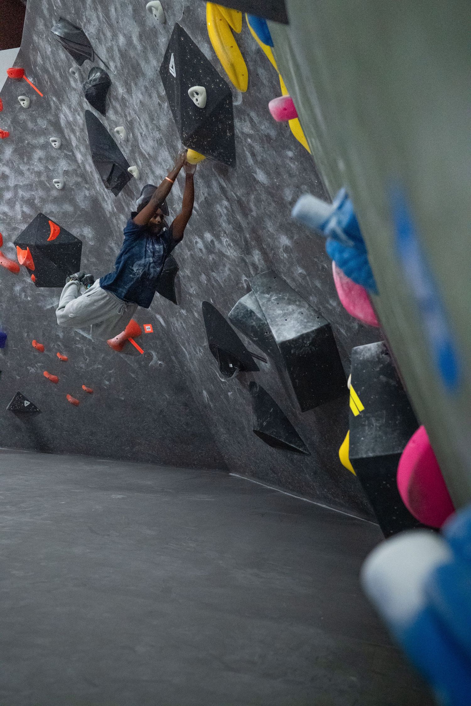 Climber in a blue shirt hangs from a black hold on an indoor bouldering wall with colorful pink, yellow and orange grips.