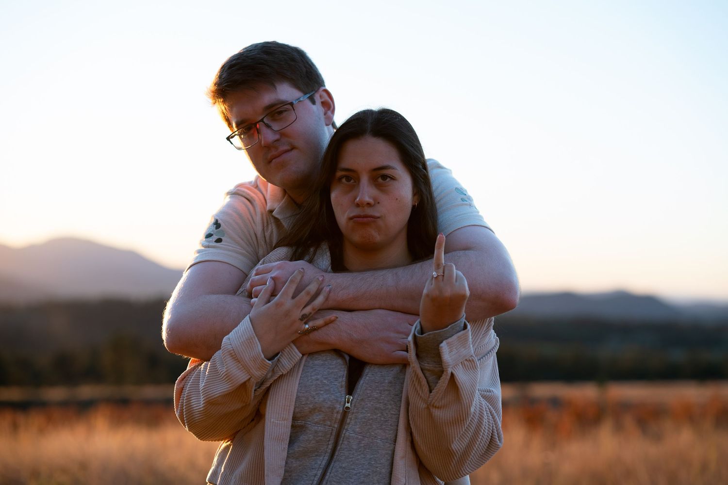 A man with glasses hugs a woman from behind at sunset in a golden field with mountains behind, as she raises a middle finger.
