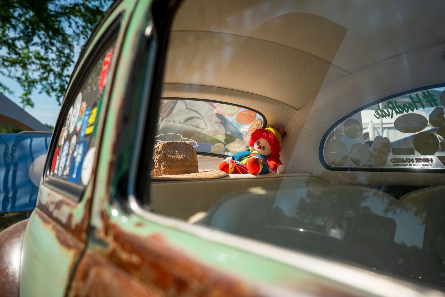 Vintage rusty car with a colorful clown doll displayed in the rear window at a car show.