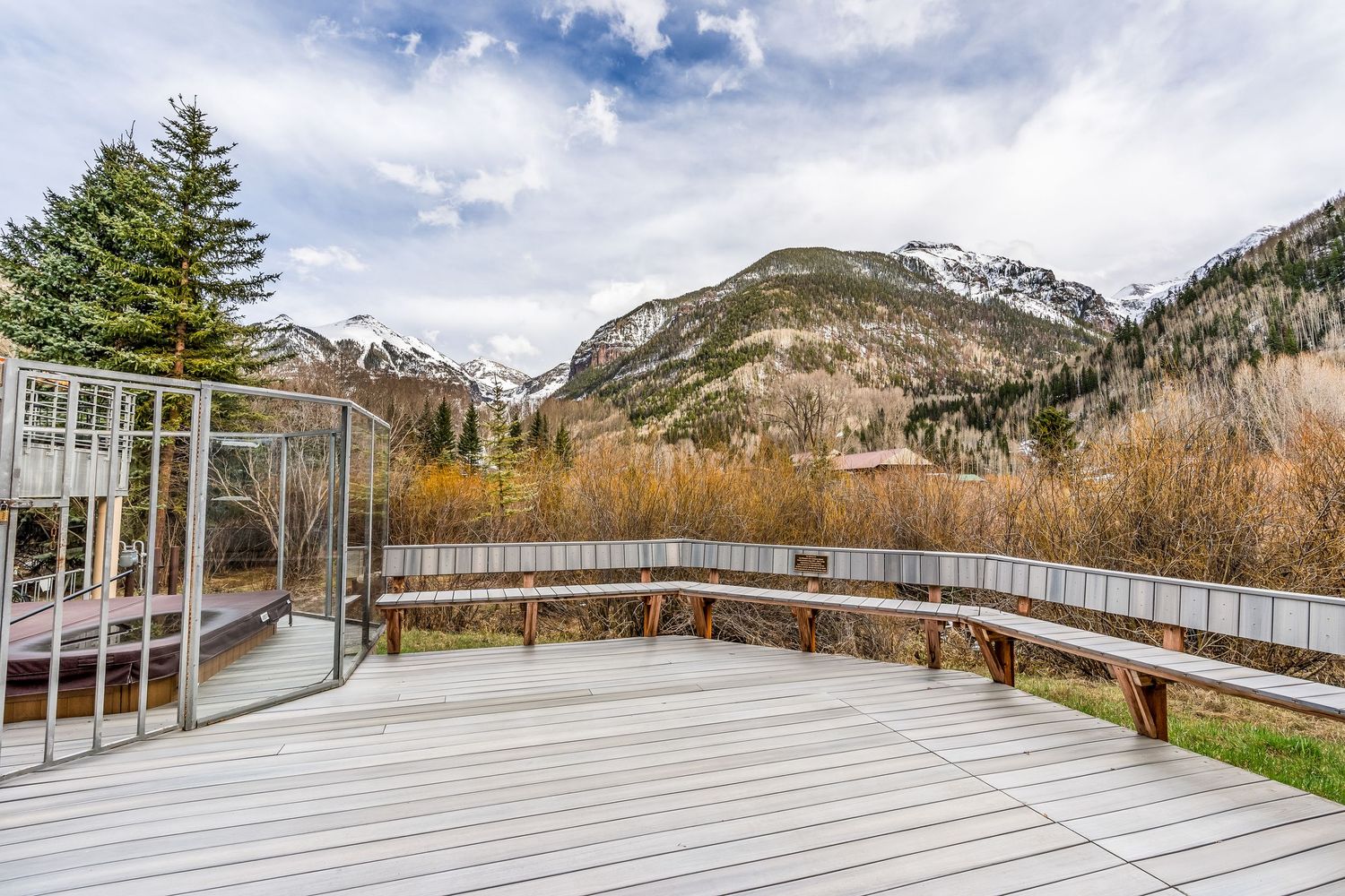 Spacious wooden deck with curved railing overlooking snow-capped mountains and autumn trees under a cloudy sky.