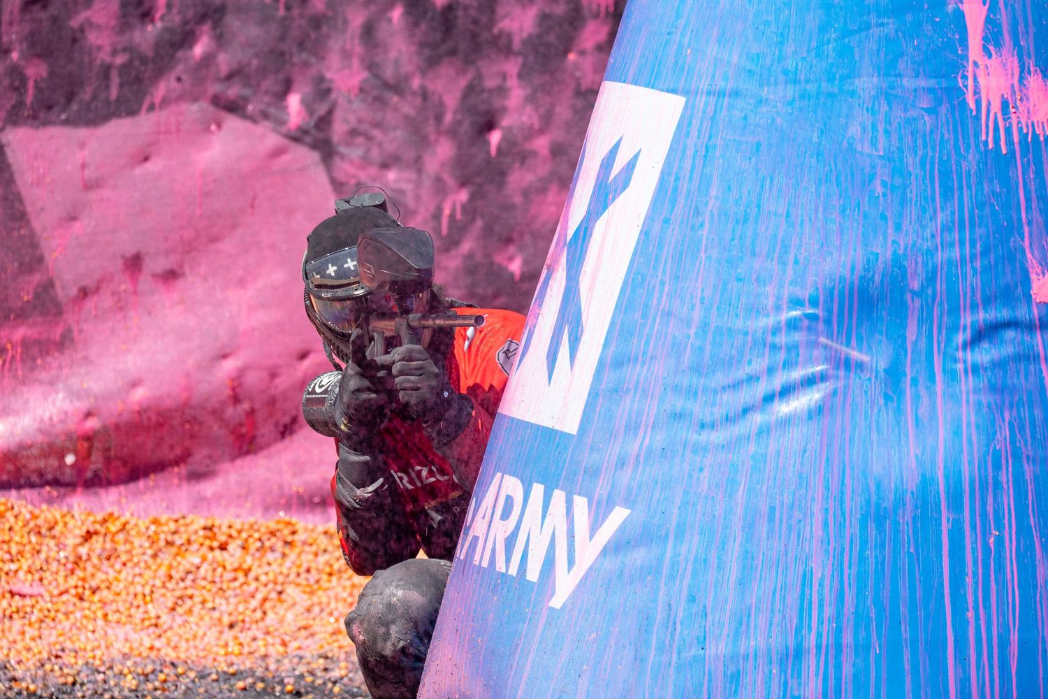Paintball player in black gear aiming marker around a blue and white Army-branded inflatable bunker.