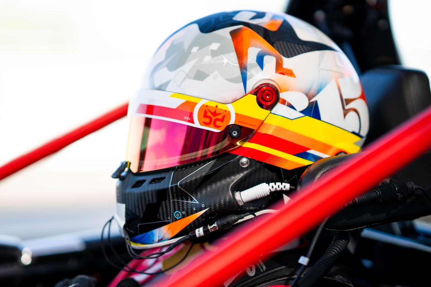 Close-up of racing helmet with blue and orange accents visible through Radical race car cockpit.
