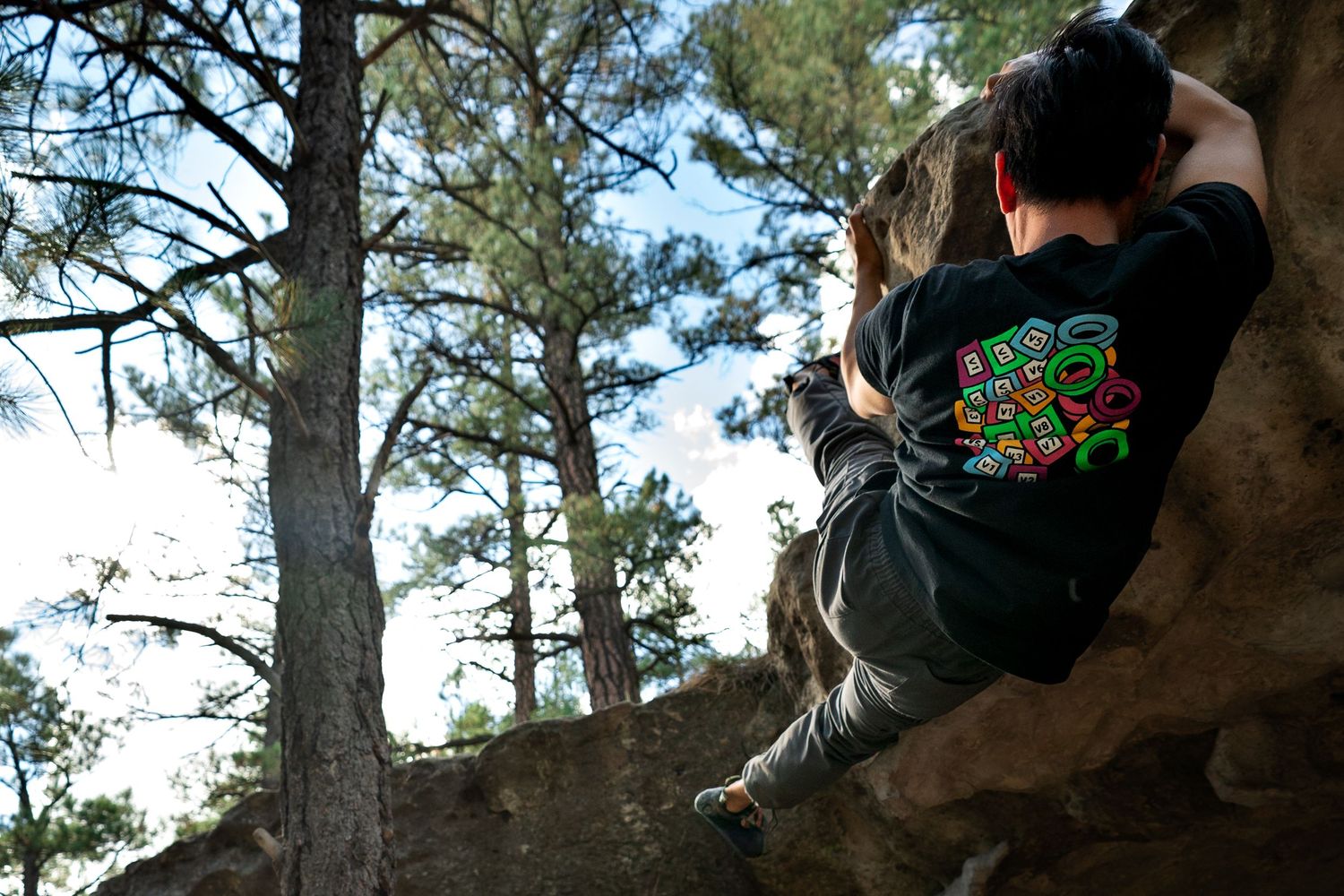 Rock climber in a black graphic t-shirt boulders up a sandstone cliff face surrounded by tall pine trees on a sunny day.