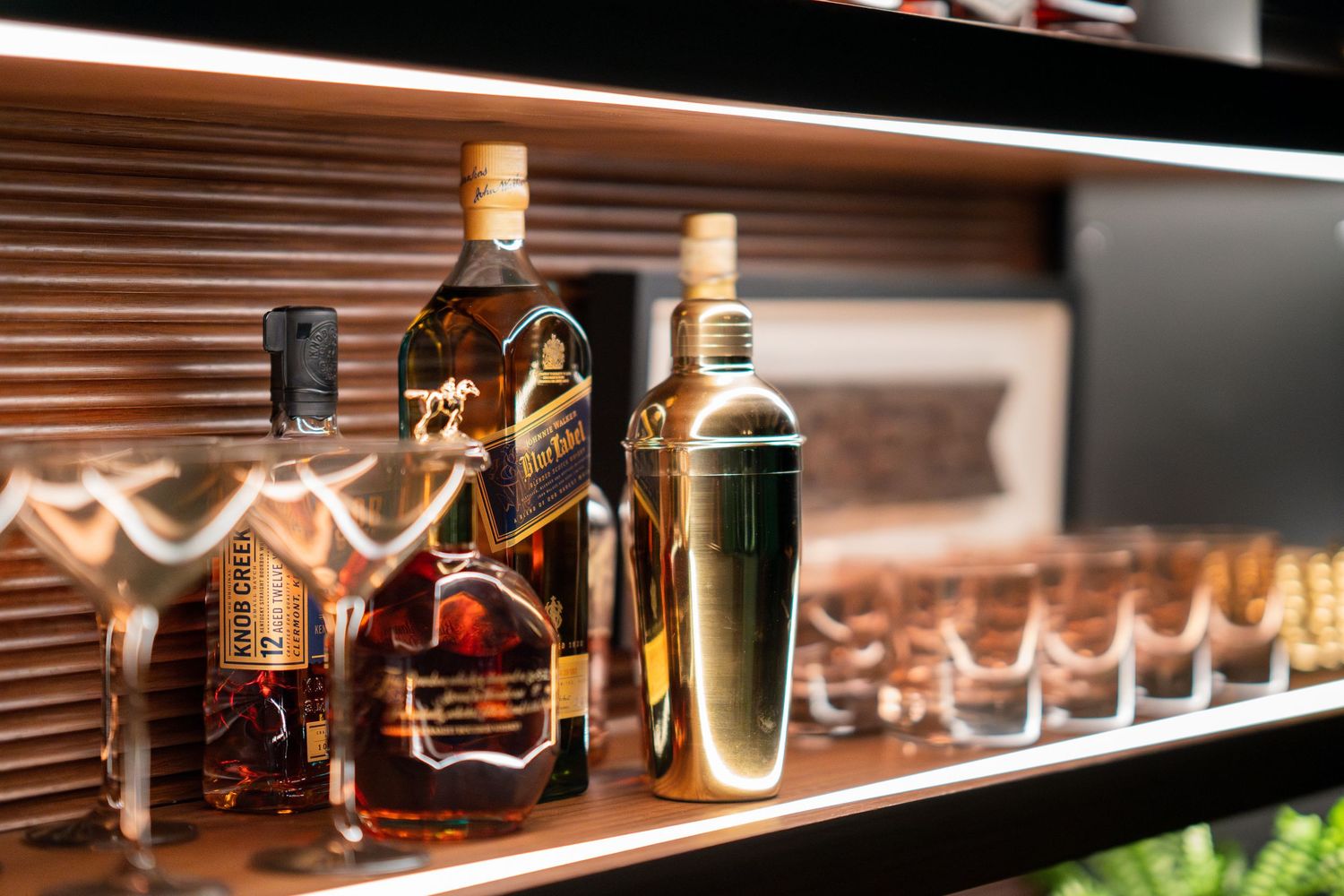 Premium whiskey bottles displayed on illuminated wooden shelf with cocktail glasses.