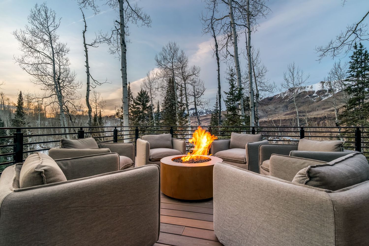 Modern mountain home deck with outdoor seating and fire pit overlooking snowy landscape at sunset.