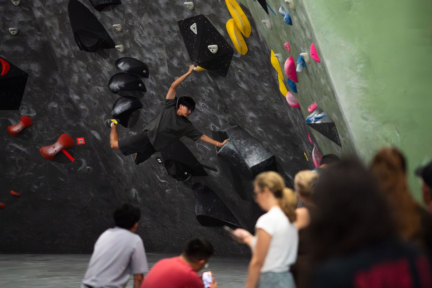 Climber in dark clothing reaches for a hold on an indoor bouldering wall with colorful holds while spectators watch from below.