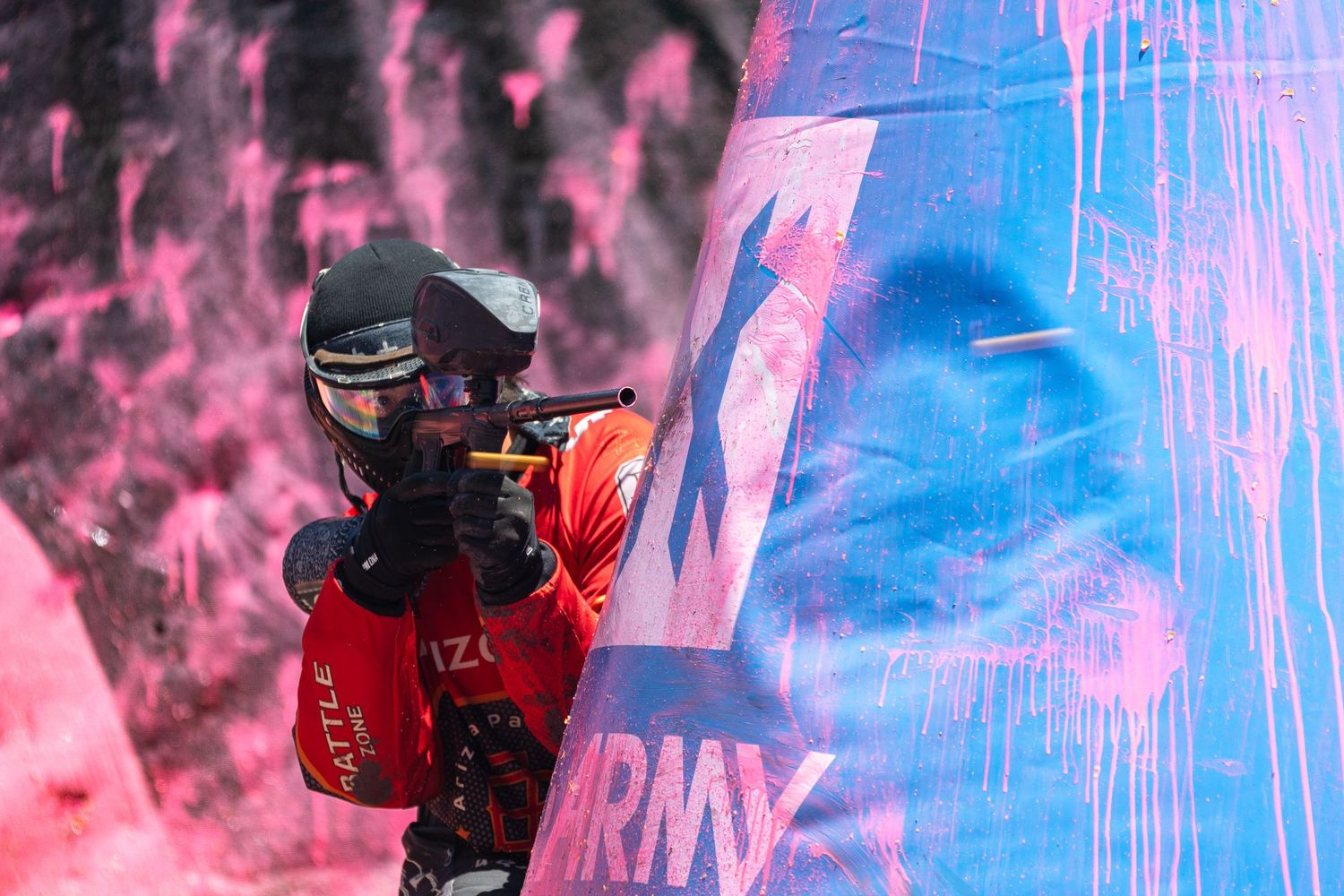 Paintball player in red gear aiming behind colorful pink and blue inflatable barrier during match.