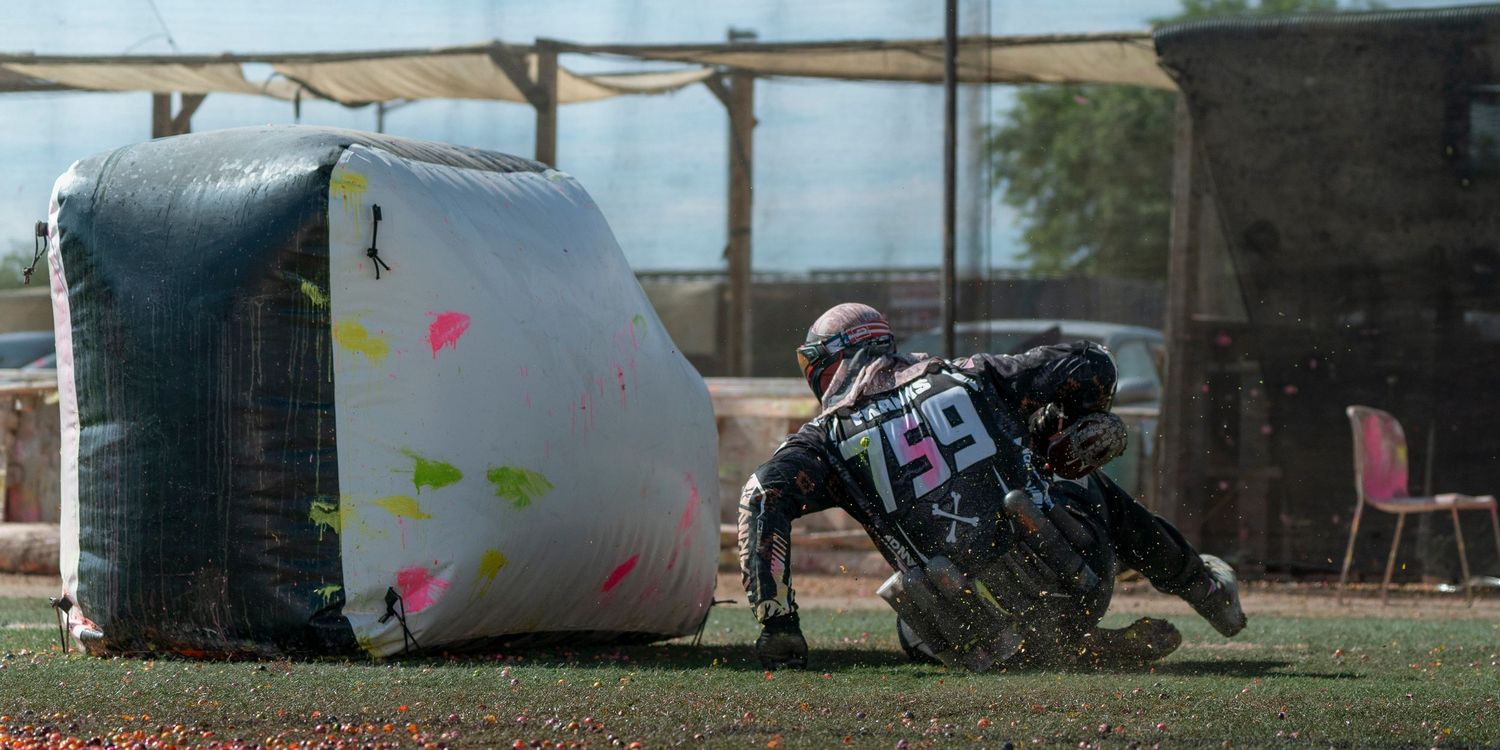 Player in tactical gear sliding behind paintball bunker during intense match.