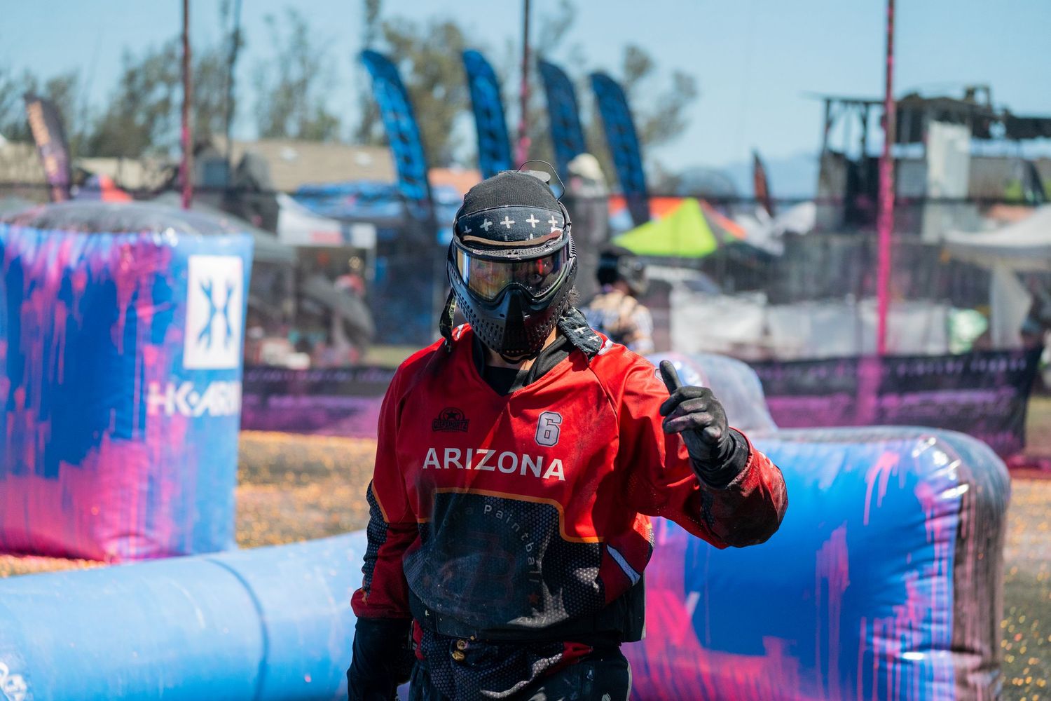 Arizona paintball player in red jersey and protective mask gives thumbs up on outdoor field.