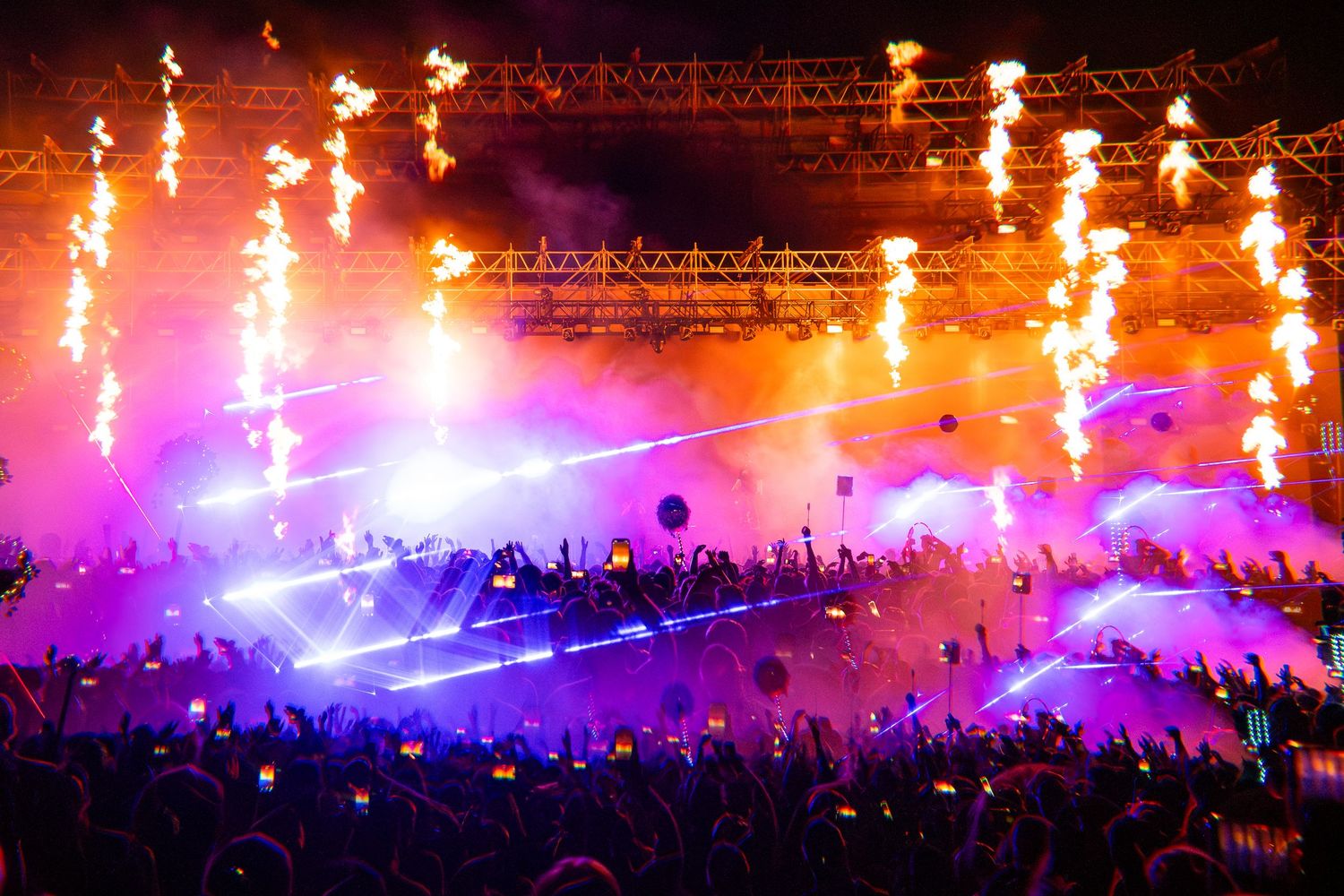 Spectacular outdoor concert stage with pyrotechnics, fire jets, and vibrant purple laser lights illuminating a massive crowd.