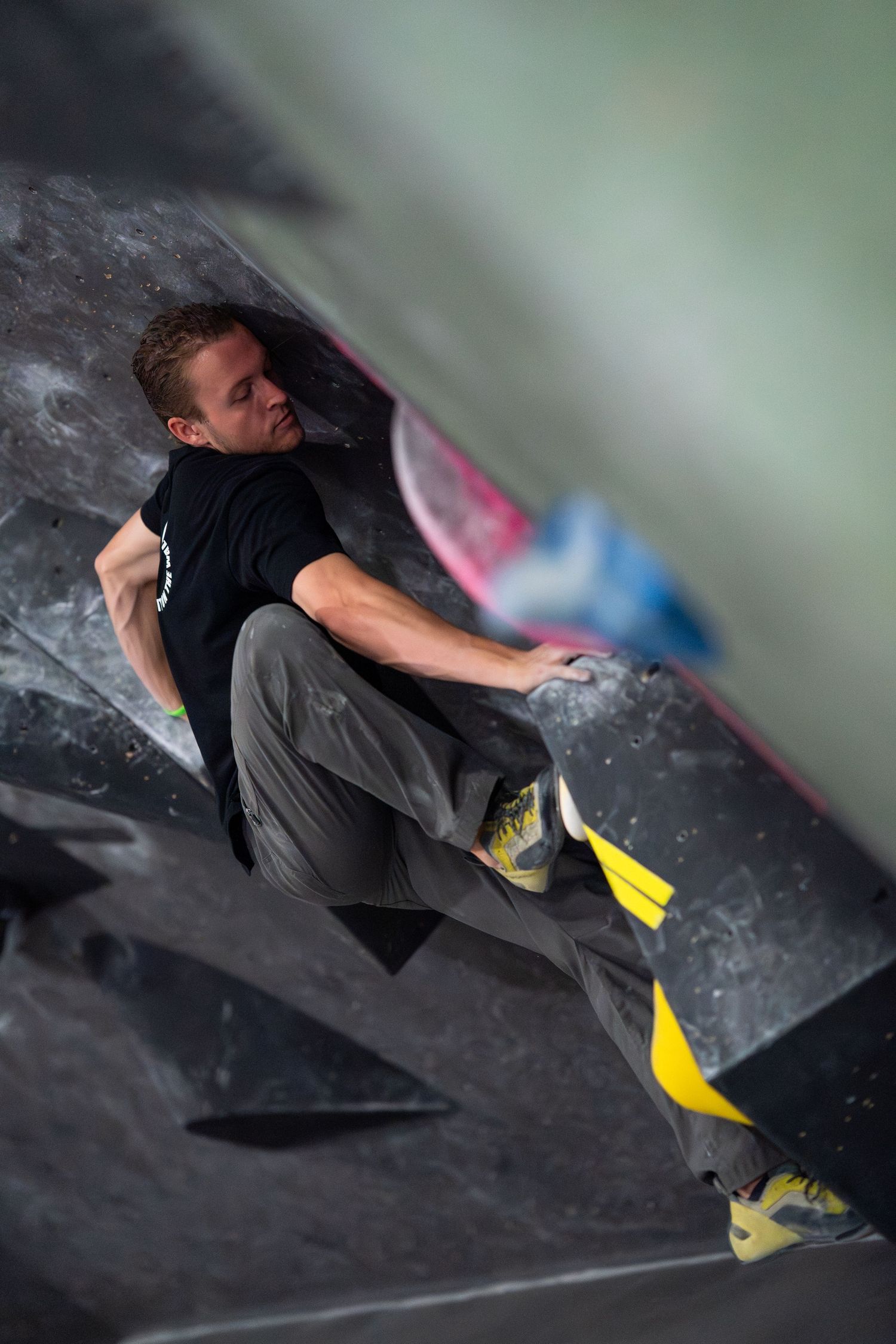 A climber in a black shirt and gray pants stretches across a dark bouldering wall, gripping a hold at an indoor climbing gym.