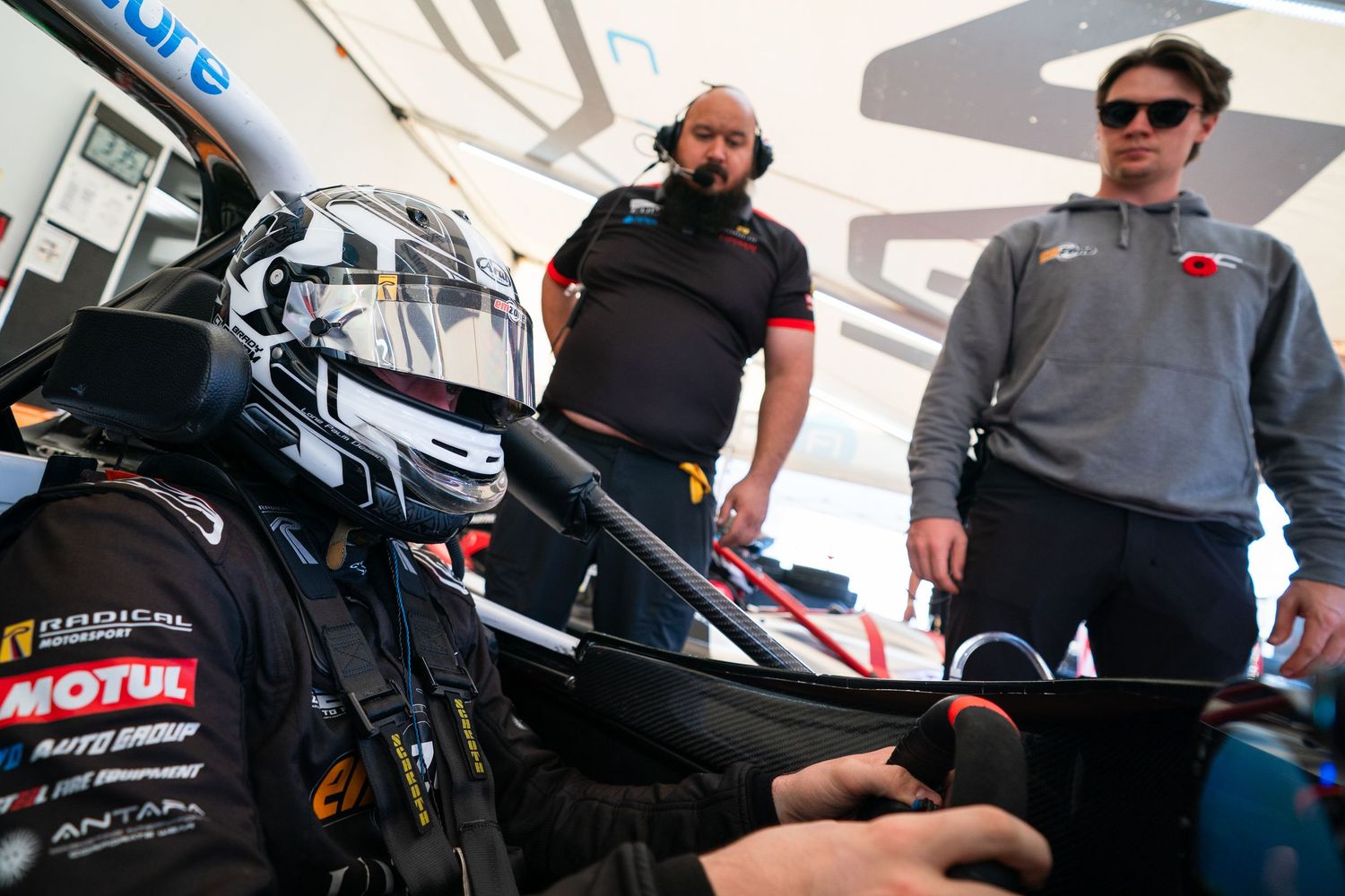 Pit crew members gather around racing vehicles under white tents during a motorsport event.