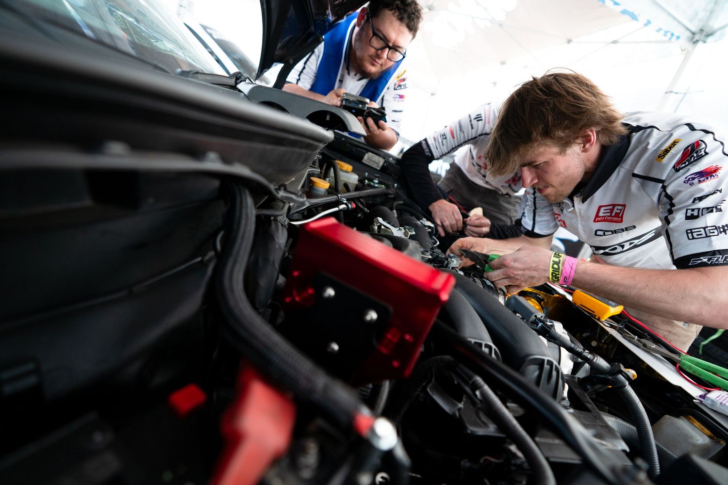Two mechanics working on a car engine in a racing pit garage under bright lights.