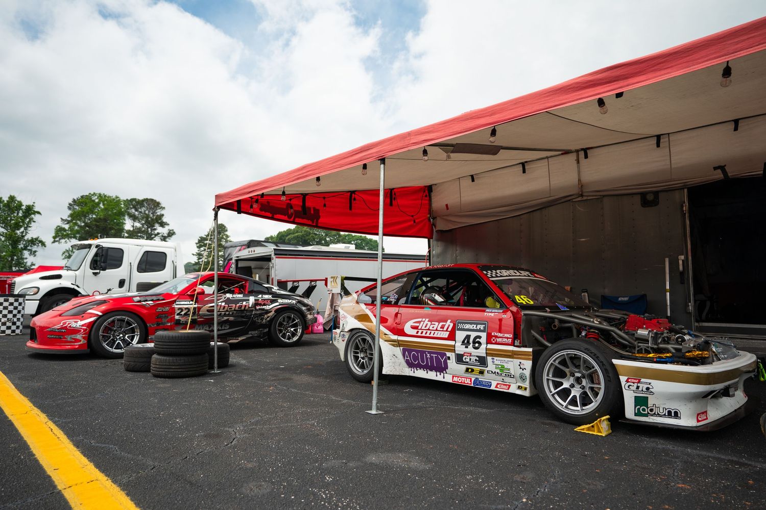 Racing cars parked under a red canopy tent in a motorsport event pit area.