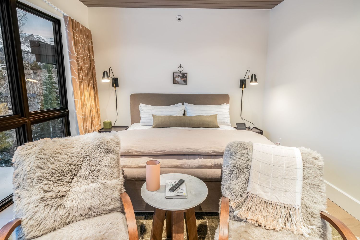 Modern bedroom with plush white bedding, cozy faux fur throws, and a small round side table near large windows.