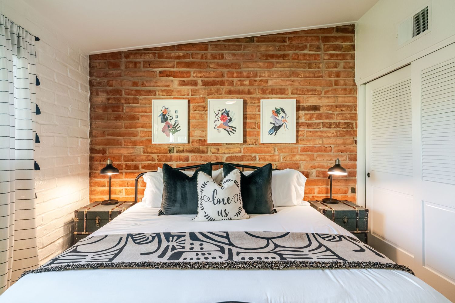 Modern bedroom interior with exposed brick wall, white bedding, and framed artwork displayed above the bed.