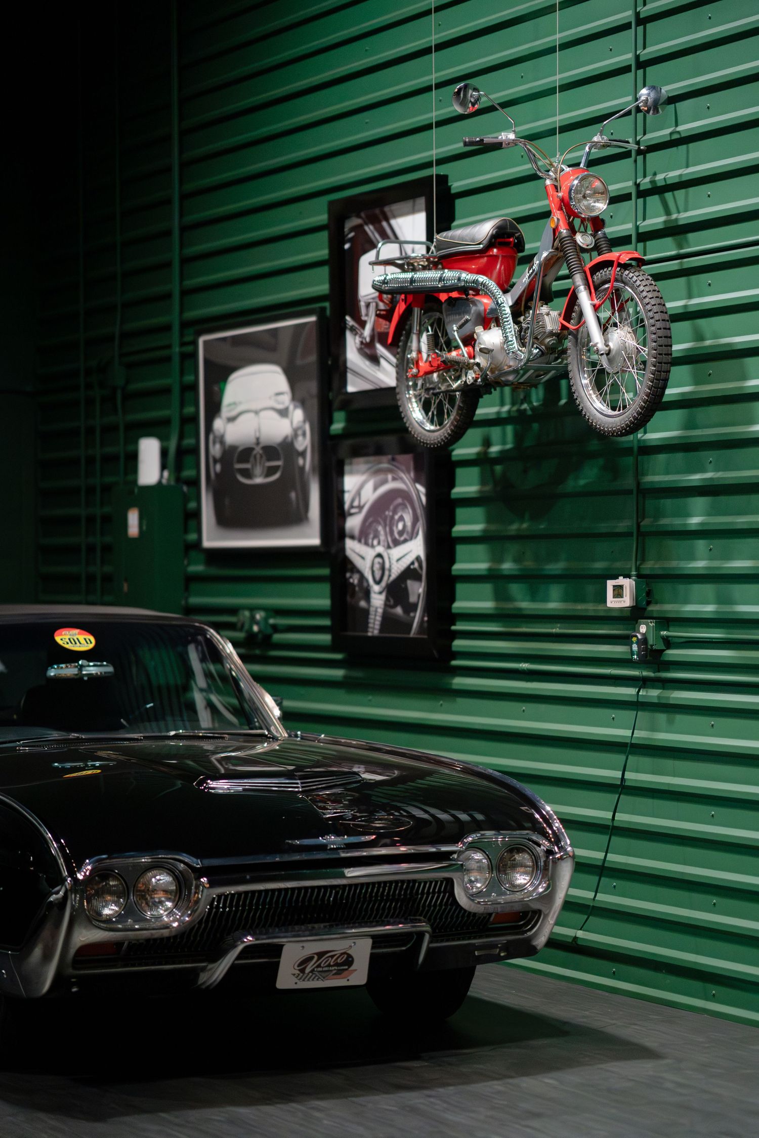 Classic black muscle car and vintage motorcycles displayed against green industrial backdrop.