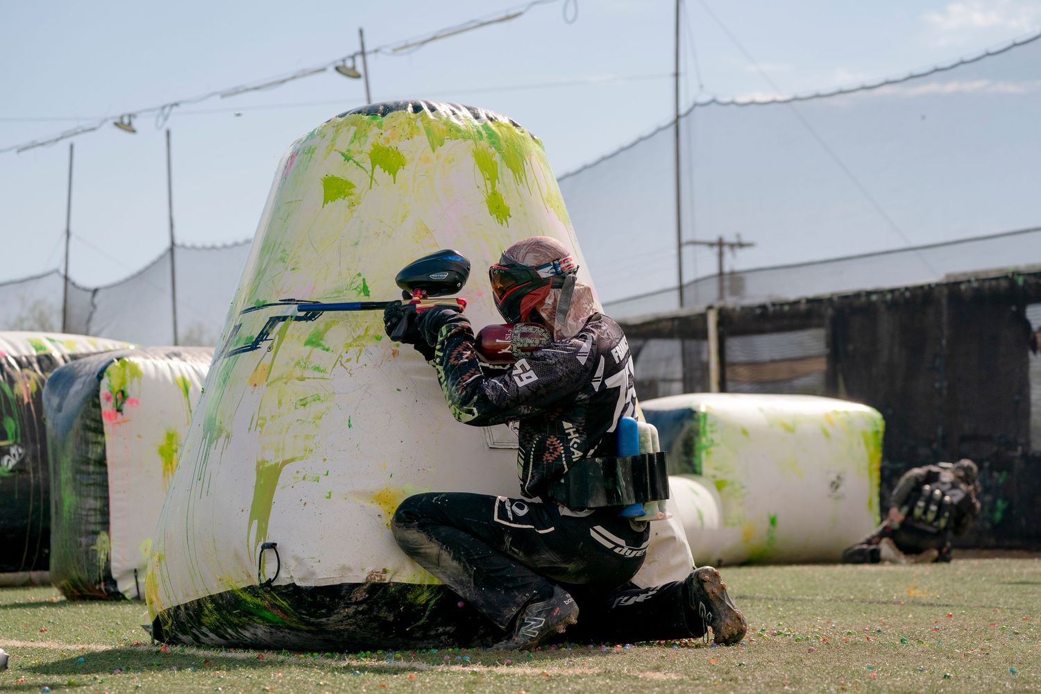 Paintball player in tactical gear crouches behind white inflatable bunker during competitive match.