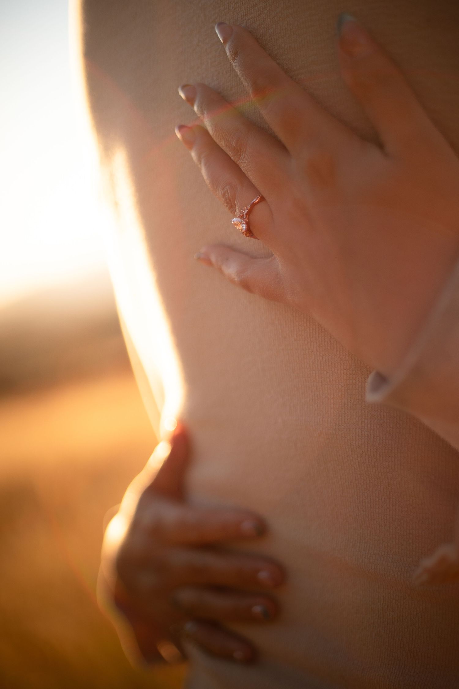 Close-up of two hands with a delicate ring resting on a person's back, softly lit by warm golden sunset light in a blurred field.