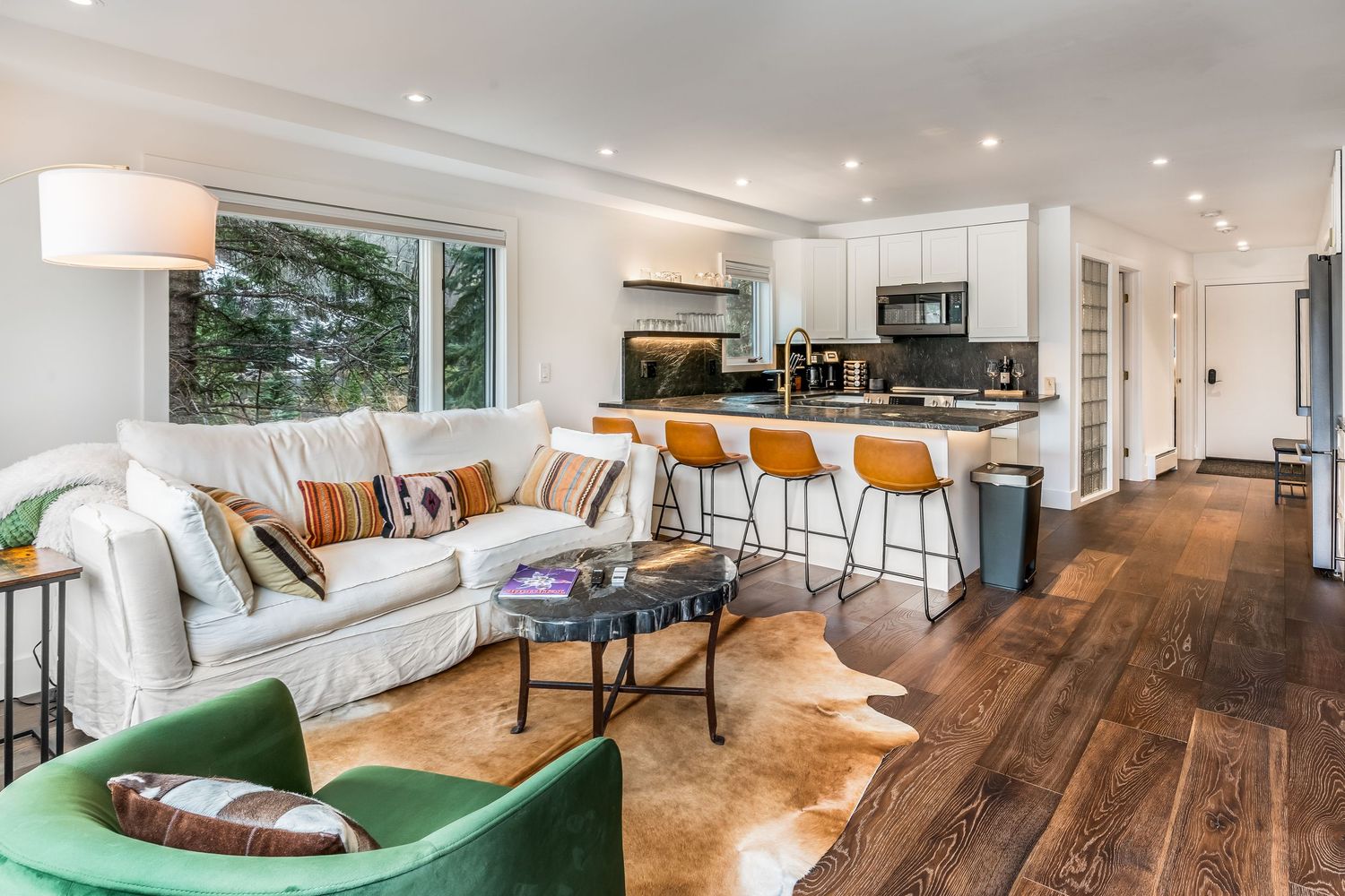 Open-concept living room with white sofa, hardwood floors, green armchair, and modern kitchen with bar stools in background.