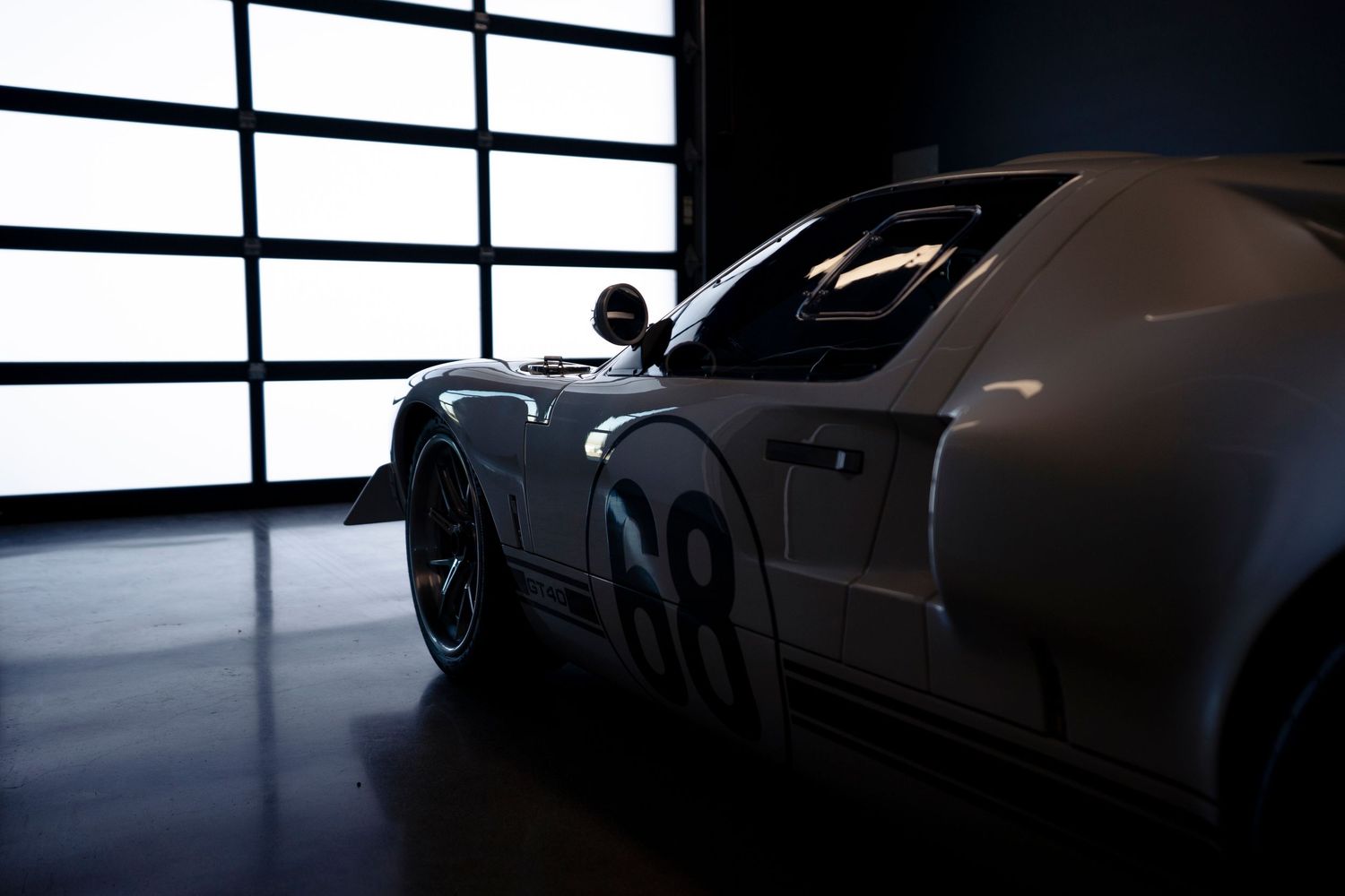 A dramatic side view of a black sports car silhouetted against bright window lighting in a dark interior space.