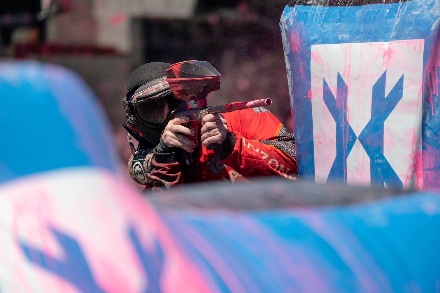 A paintball player in red gear aims their marker behind a colorful inflatable bunker during a competitive match.