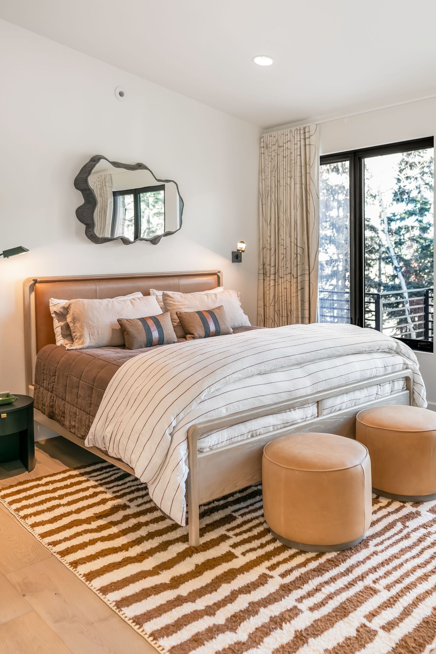 Modern bedroom with striped area rug features neutral decor, large windows, and round leather ottomans.