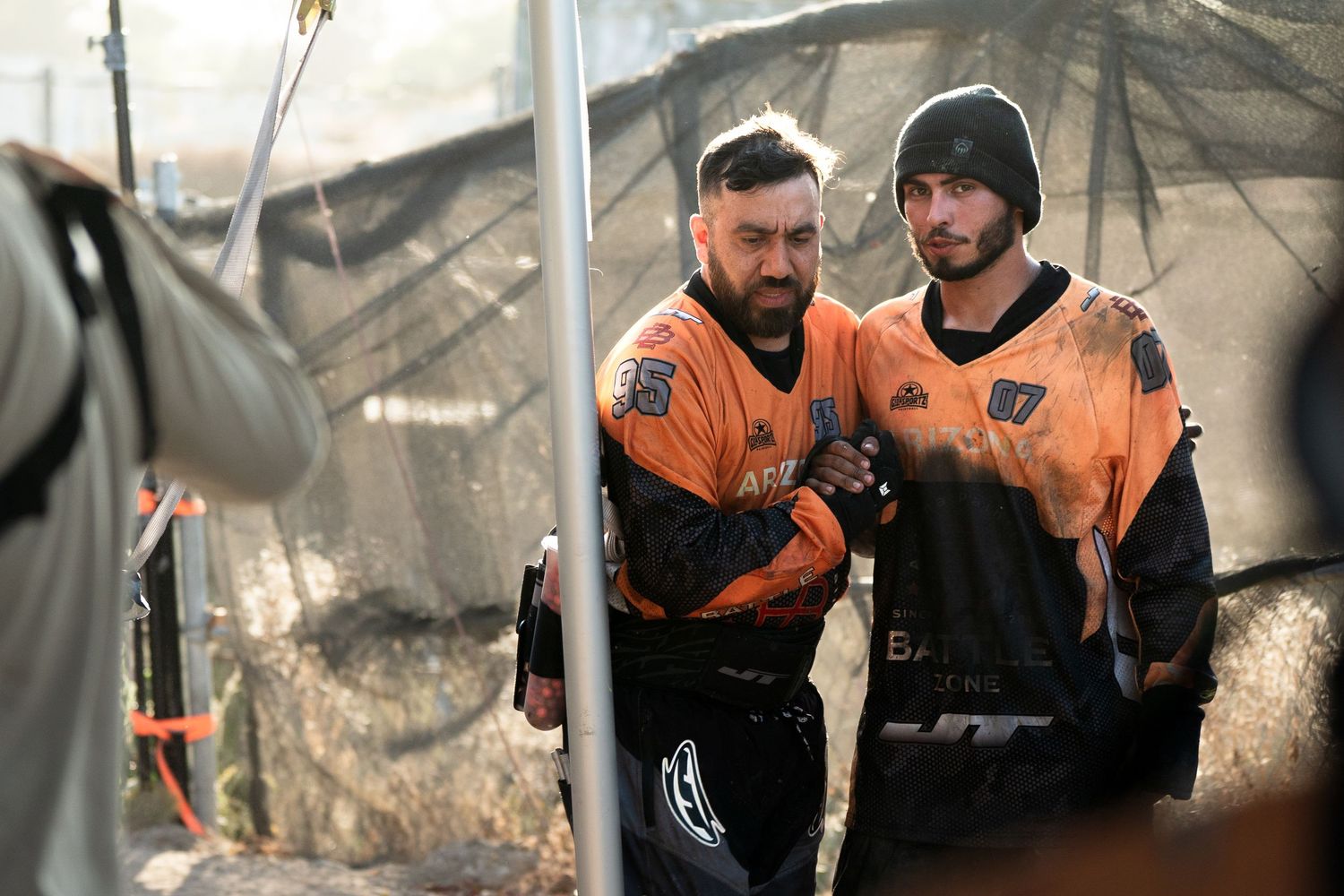 Two paintball players in orange and black jerseys strategizing together during an outdoor paintball tournament.