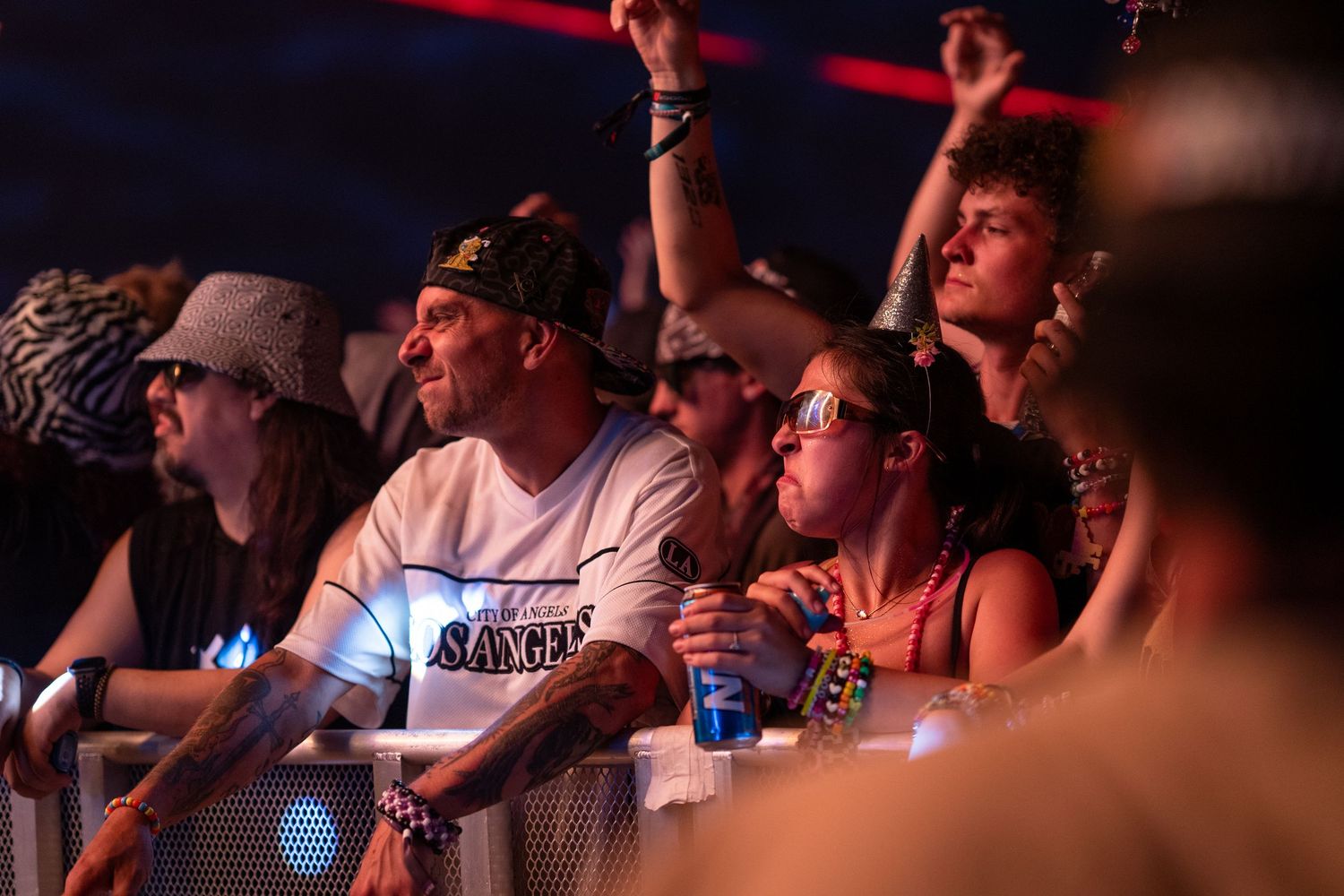 Crowd of festival-goers watching a live concert at night, wearing sunglasses and casual attire near the stage barrier.