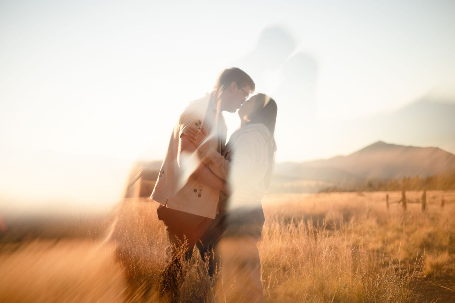 Couple embracing in a golden grassy field at sunset with soft double-exposure glow and rolling mountains in the background.