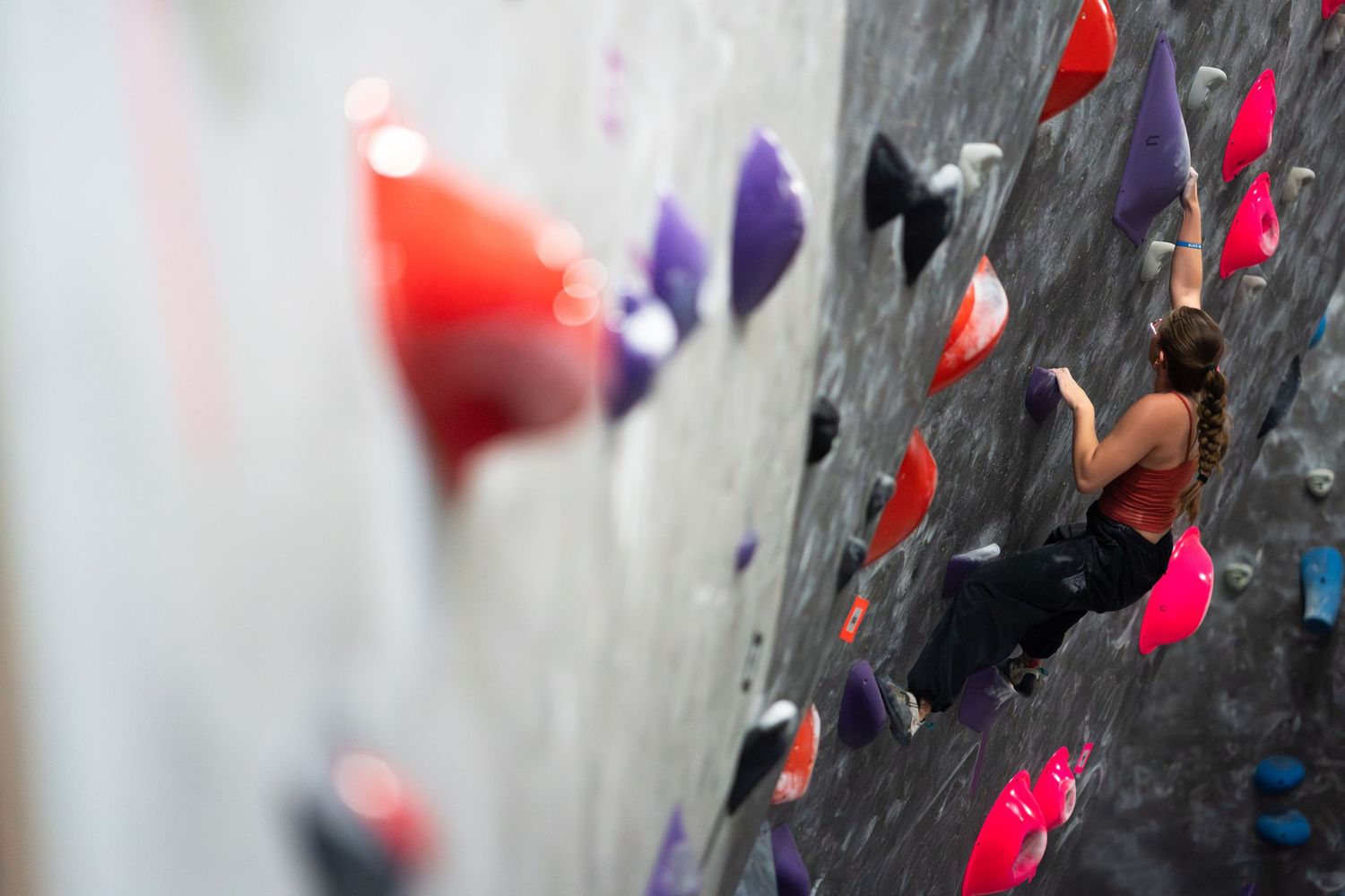 Climber in a red top and dark pants reaches for a hold while scaling a colorful indoor bouldering wall with red, pink and purple grips.