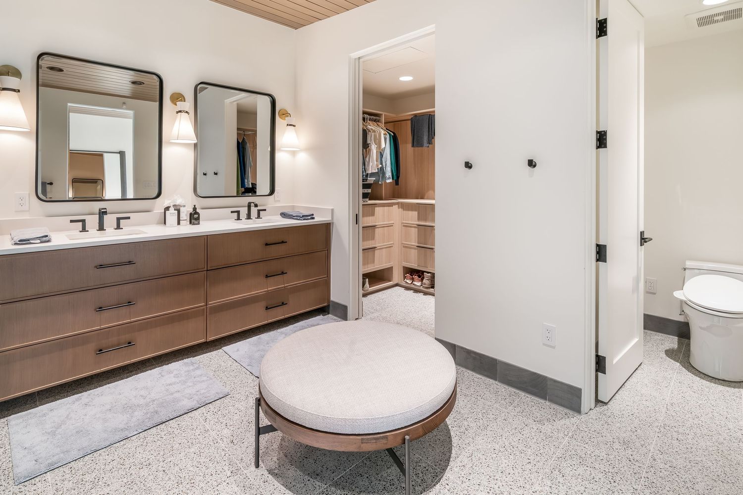 Contemporary bathroom with double vanity, round ottoman, and wood accents.