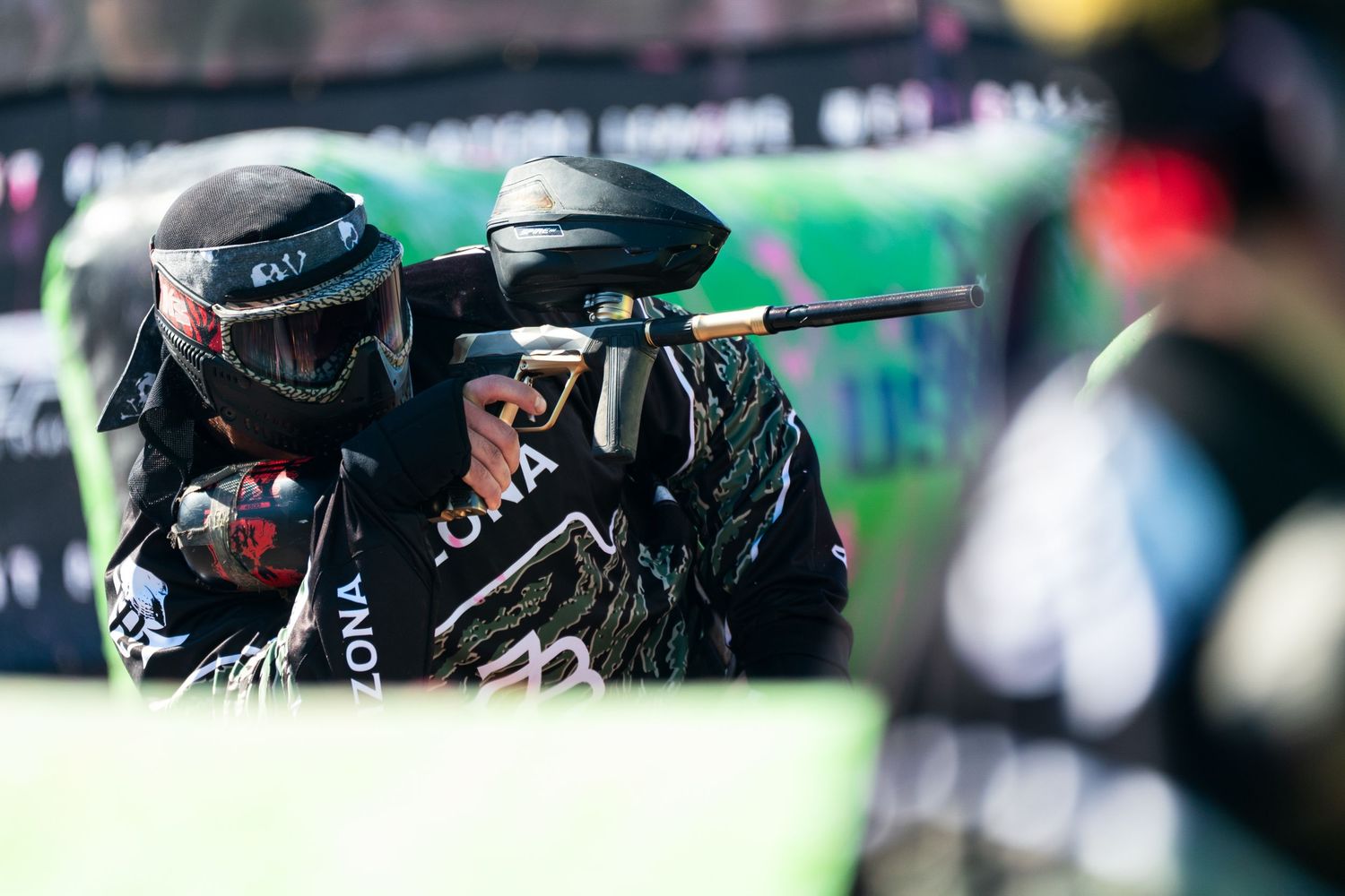 Paintball player in tactical gear aiming marker during competitive tournament match outdoors.