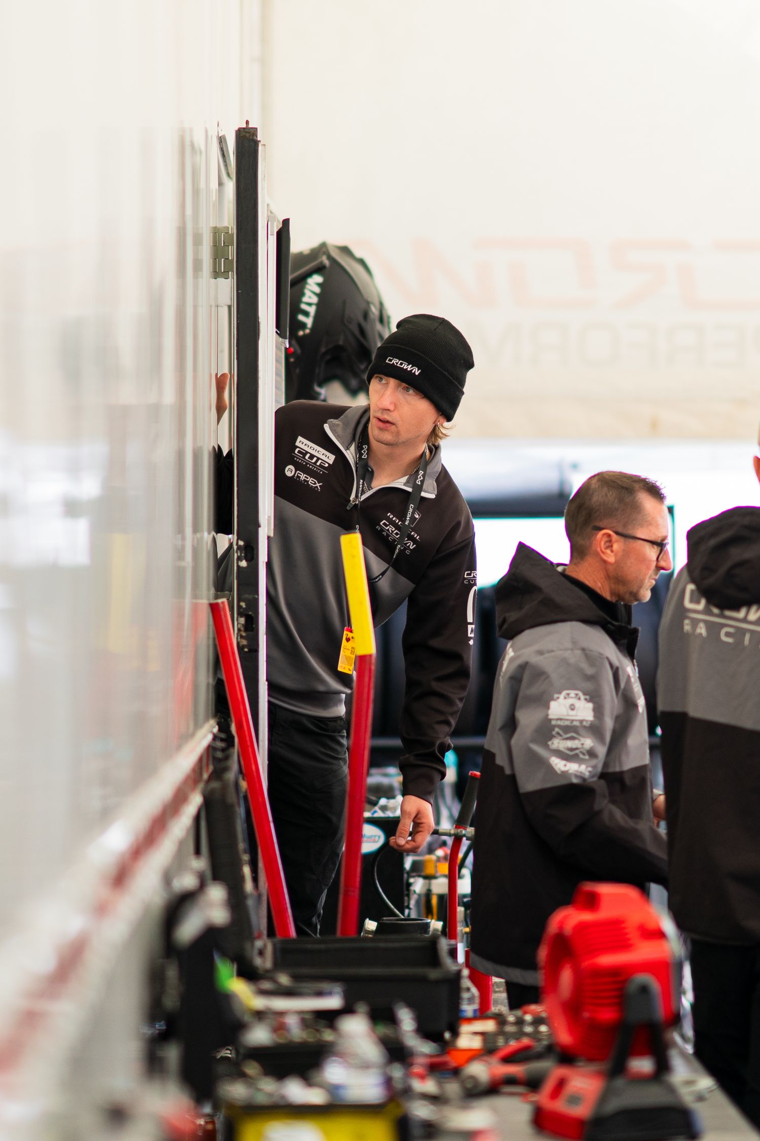 Racing team mechanics work under a tent canopy making adjustments to equipment during a motorsport event.