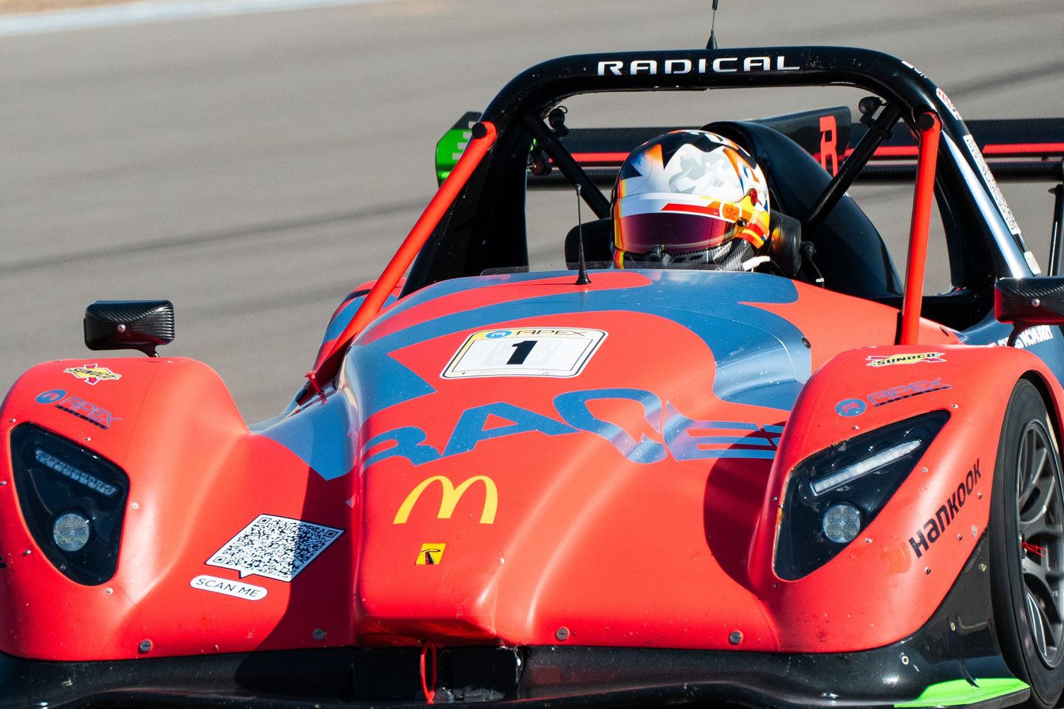 Red Radical sports car on racetrack with driver visible through windscreen.