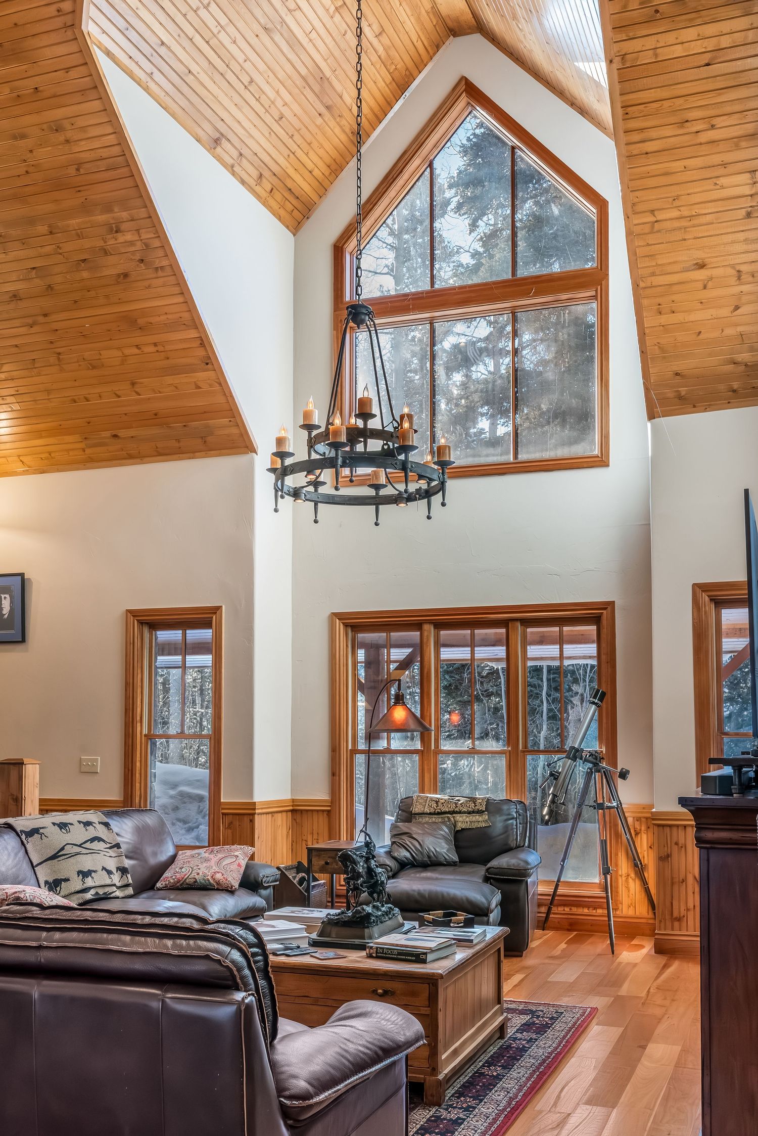 Rustic living room with vaulted ceiling, large windows, wood beams, and a wrought iron chandelier.