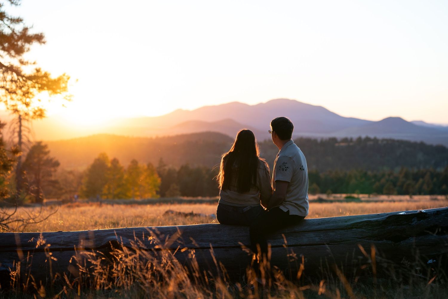 Couple sits close together on a rustic wooden fence, watching a golden sunset glow over distant mountains and a forested valley.